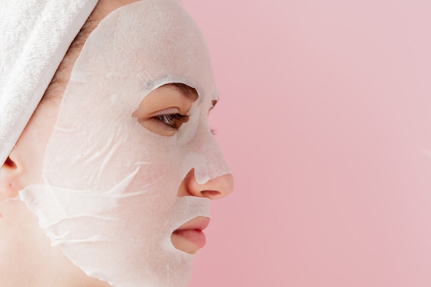 La mujer joven hermosa está aplicando una máscara cosmética del tejido en una cara en un fondo rosado. Concepto de tecnología y tratamiento de salud y belleza.