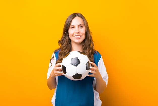 La mujer joven hermosa está sosteniendo un balón de fútbol sobre fondo amarillo.