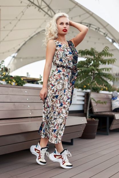 Mujer joven hermosa con estilo en un vestido de moda con un patrón y zapatillas de deporte en la ciudad