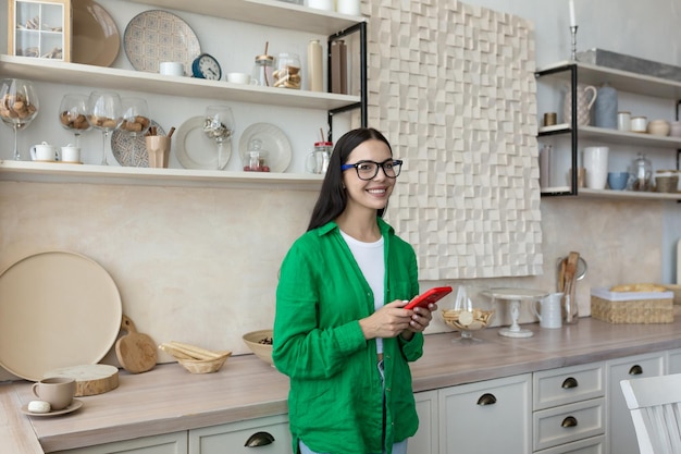 Una mujer joven y hermosa con gafas y una camisa verde está parada en la cocina escribiendo un