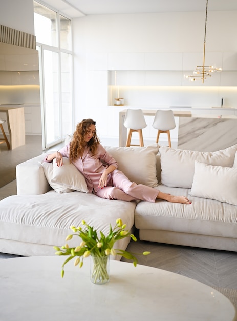 Mujer joven hermosa con el pelo rizado en pijama rosado que miente en el sofá blanco del sofá en la mañana. Salón de estilo escandinavo y cocina interior.