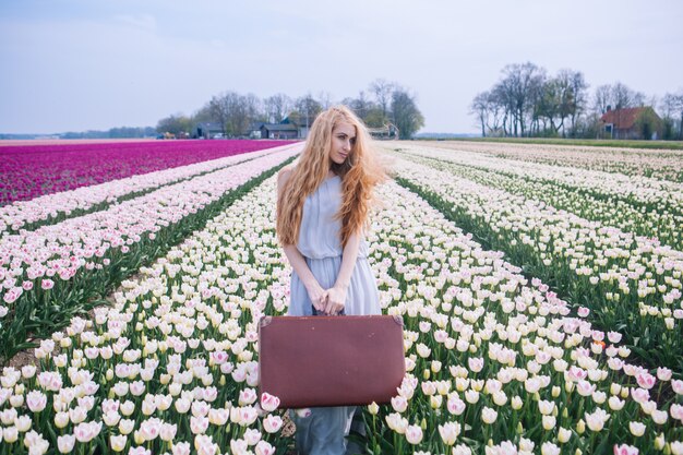 Mujer joven hermosa con el pelo rojo largo que lleva en el vestido blanco que se coloca con la maleta vieja del vintage en campo colorido del tulipán.