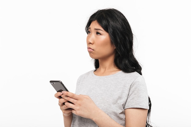 mujer joven hermosa pensativa asiática que presenta aislada sobre la pared blanca que usa el teléfono móvil.