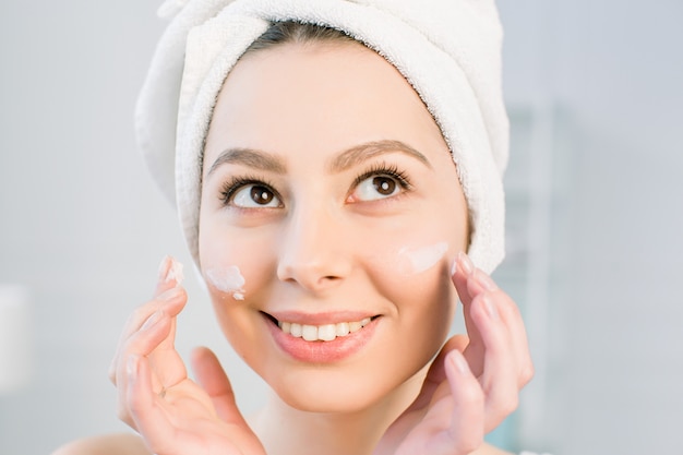 Mujer joven hermosa que aplica la crema en su cara en baño. Tratamiento para un concepto de piel perfecto.