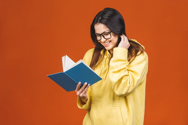 Foto mujer joven hermosa que lleva la situación casual aislada sobre el fondo anaranjado, leyendo un libro.