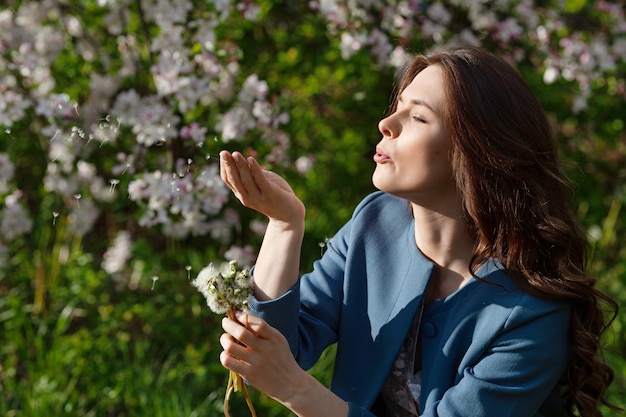 Mujer joven hermosa que sopla el diente de león en el jardín de primavera. Disfruta de la naturaleza. Niña sonriente sana al aire libre. Concepto libre de alergias. Libertad