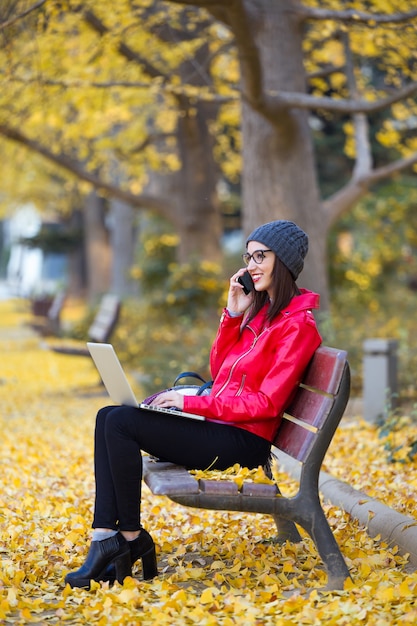 Mujer joven hermosa que usa su teléfono móvil mientras que trabaja con la computadora portátil en otoño.