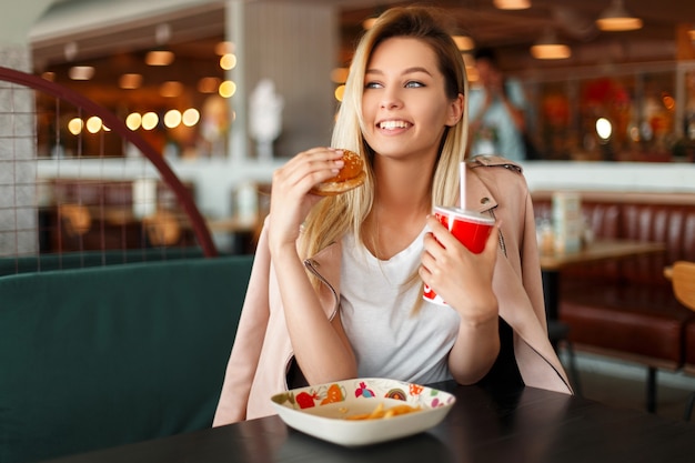 Mujer joven hermosa satisfecha con una sonrisa comiendo una hamburguesa y bebiendo cola en el interior