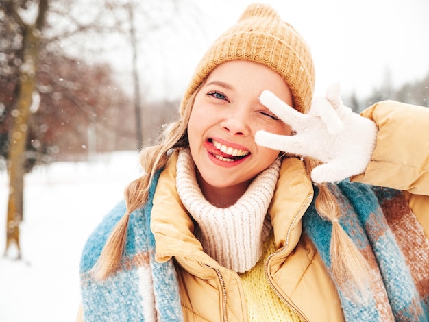 Mujer joven hermosa sonriente hipster en ropa de abrigo de moda y bufanda. Mujer despreocupada posando en la calle en el parque. Modelo puro positivo divirtiéndose en la nieve. Disfrutando de los momentos invernales. Muestra el signo de la paz