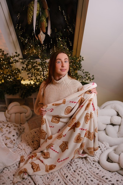 Mujer joven hermosa y sonriente se queda en la cama con el hombre de la tarta en envoltura con suéter blanco en la habitación decorada para celebrar el año nuevo y el estado de ánimo festivo de Navidad