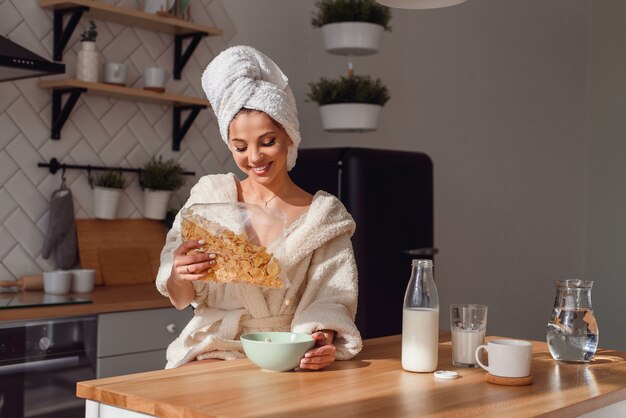 La mujer joven hermosa vierte las avenas en placa con leche. La niña tiene un desayuno saludable en la acogedora cocina con estilo en la mañana.