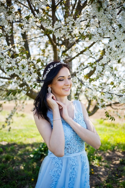 Mujer joven con hermoso maquillaje con un aro vintage en un vestido azul claro con un ramo posando sobre un fondo de follaje verde en un parque. Chica atractiva retrato fresco con flores blancas.