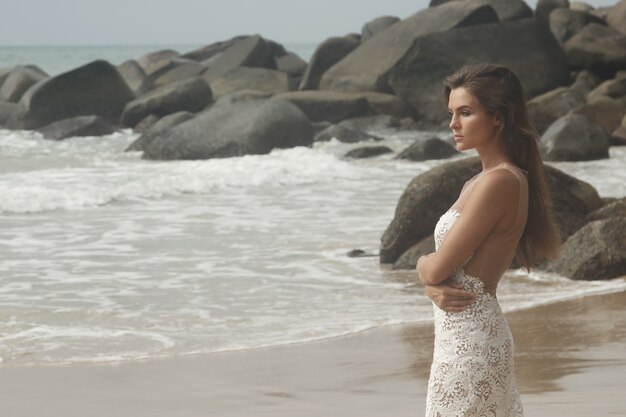 Mujer joven con hermoso vestido blanco está posando en la playa rocosa