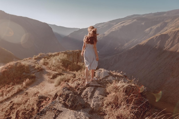 Una mujer joven con un hermoso vestido se encuentra al borde de una colina y mira la soleada vista de la montaña de verano