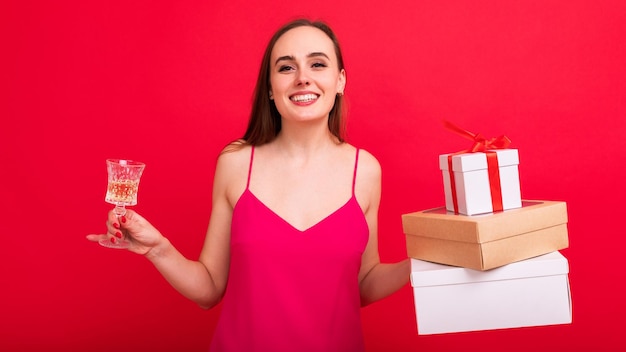 Una mujer joven con un hermoso vestido sostiene una copa de champán y regalos para Navidad Una modelo posa en el estudio sobre un fondo rojo