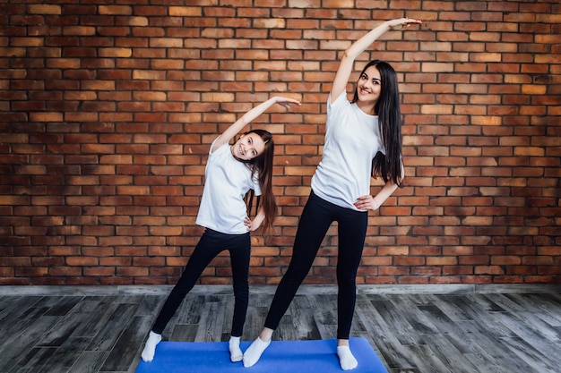 Mujer joven con hija en ropa deportiva practicando yoga en casa