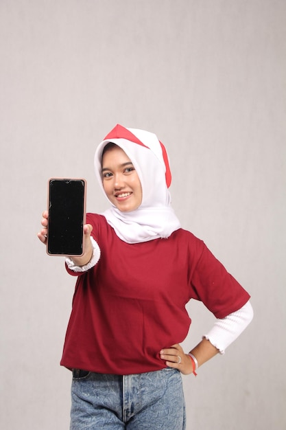 mujer joven en hijab en camisa roja que muestra el teléfono móvil de pantalla negra a la cámara con hermosos dientes s
