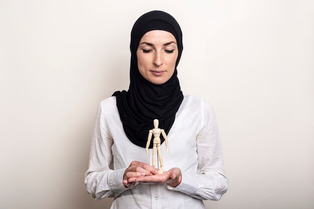 Una mujer joven con un hijab está sosteniendo mira una marioneta de un hombre de madera sobre un fondo claro.