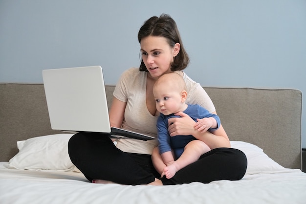 Mujer joven con hijo pequeño sentado en la cama con ordenador portátil, madre freelance, coach, blogger que trabaja en casa