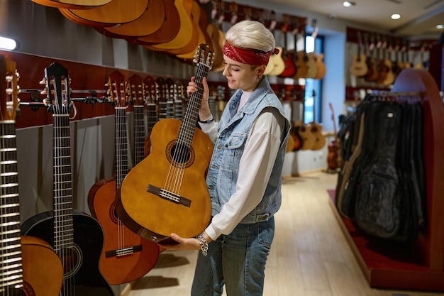 Mujer joven hipster eligiendo guitarra acústica en la tienda de instrumentos. Compradora femenina comprando en una tienda musical