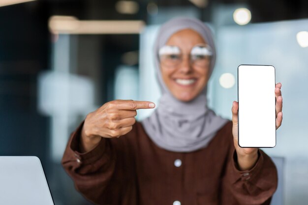 Una mujer joven en un hiyab en un fondo borroso sostiene un teléfono en sus manos sonriendo muestra un