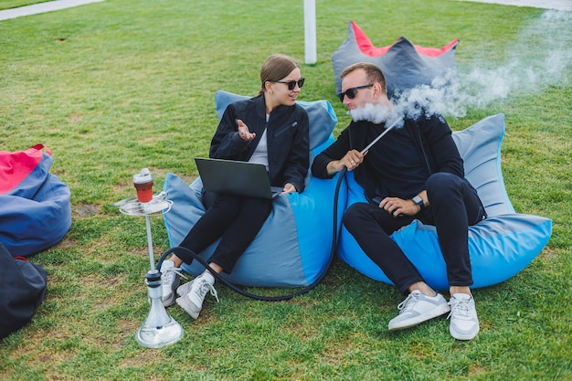 Una mujer joven y un hombre están sentados en un parque en una bolsa de frijoles y fumando narguile Pareja feliz relajándose al aire libre con narguile