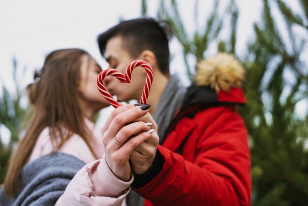 Una mujer joven y un hombre sostienen bastones de caramelo en forma de corazón y se besan contra el árbol de Navidad