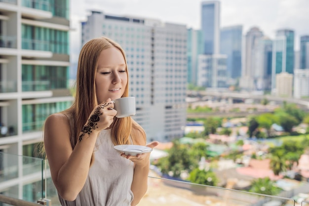 Mujer joven con una imagen en la mano de henna - mehendi bebe café por la mañana en el balcón con vistas a la gran ciudad y los rascacielos