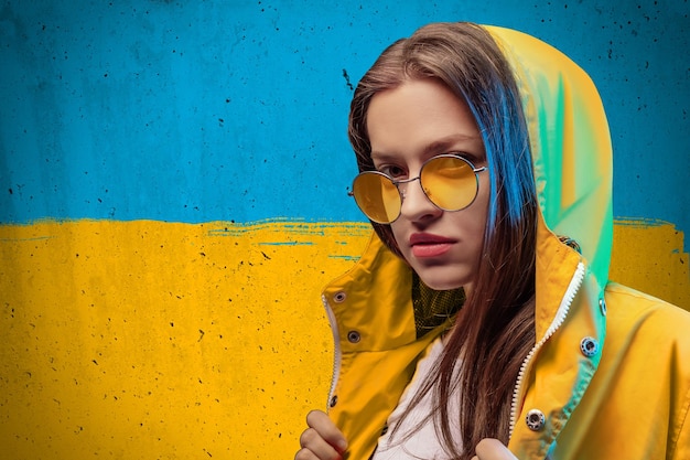 Mujer joven con impermeable amarillo y gafas de color redondas en el fondo de la bandera ucraniana