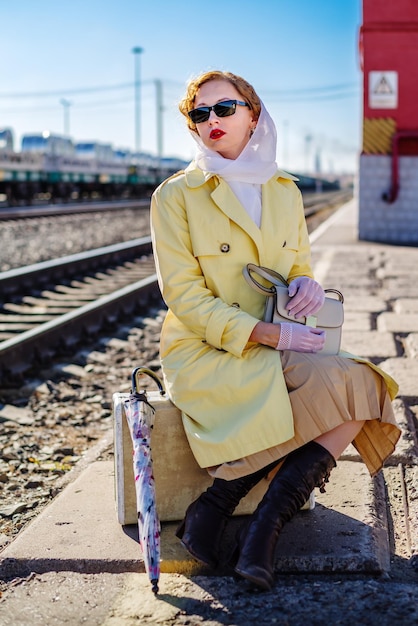 Mujer joven en impermeable amarillo y gafas oscuras se sienta en la maleta en el andén de la estación de tren