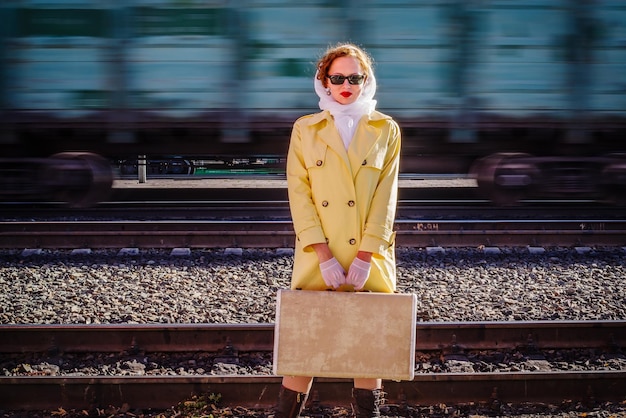 Una mujer joven en impermeable amarillo con maleta en sus manos se encuentra en el andén de la estación de tren
