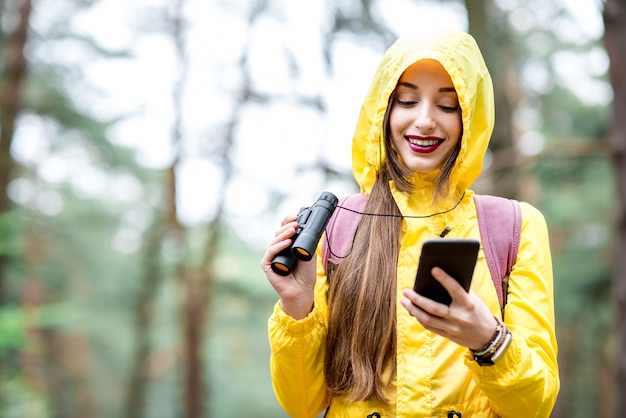 Mujer joven en impermeable amarillo con teléfono inteligente para orientación durante la caminata en el bosque de pinos verdes