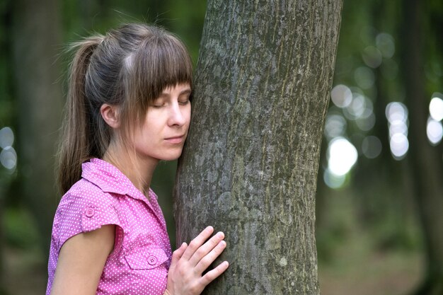 Mujer joven inclinada al tronco de un árbol abrazándolo con sus manos en el bosque de verano.