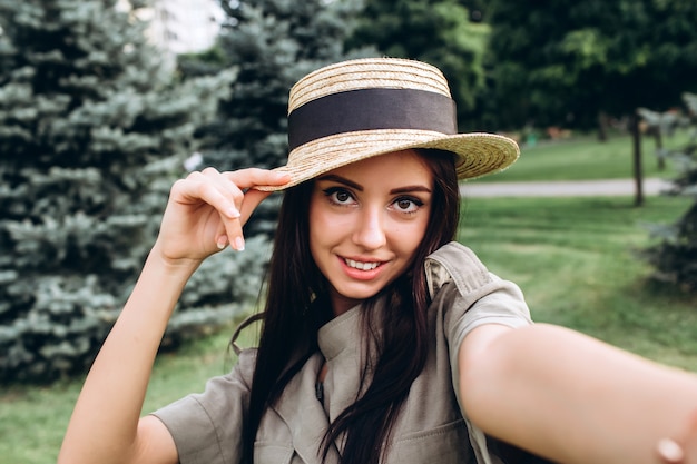 Mujer joven inconformista morena tomando un selfie, mostrando la lengua, cara divertida posando al aire libre. Closeup retrato de una niña en un elegante sombrero de verano en un parque.