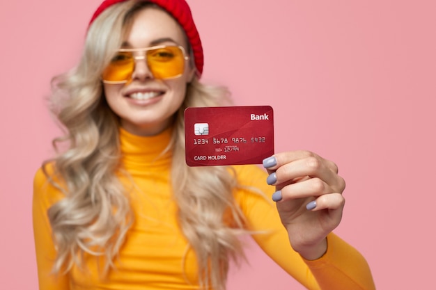 Mujer joven inconformista optimista con gorro de punto y gafas de sol que demuestran la tarjeta bancaria mientras hacen publicidad del servicio bancario contra el fondo rosa