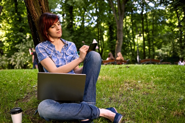 Mujer joven independiente hermosa que usa la computadora portátil que se sienta debajo del árbol en el parque.