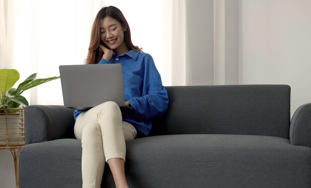 Mujer joven independiente de negocios asiática trabajando en una computadora portátil revisando las redes sociales mientras se acuesta en el sofá cuando se relaja en la sala de estar en casa