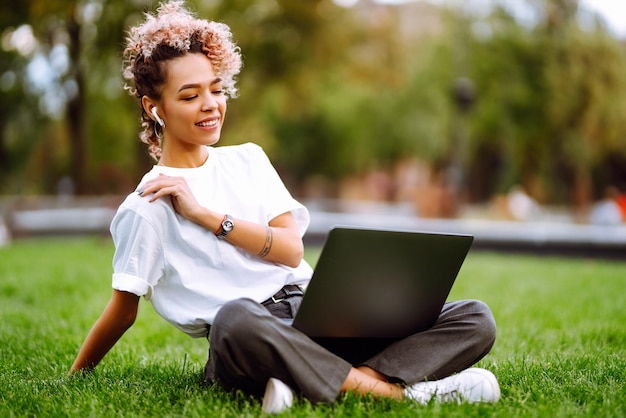 Mujer joven independiente que disfruta viendo un seminario web educativo en una computadora portátil sentada en la hierba verde