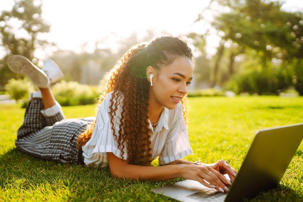 Mujer joven independiente sentada en la hierba verde con una computadora portátil Educación en línea