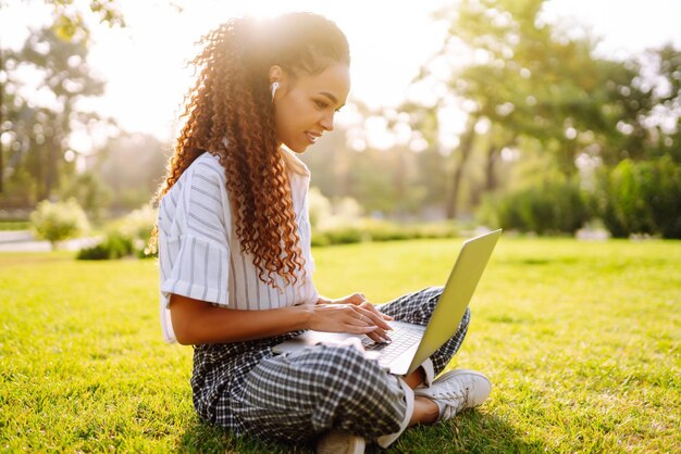 Mujer joven independiente sentada en la hierba verde con una computadora portátil Educación en línea