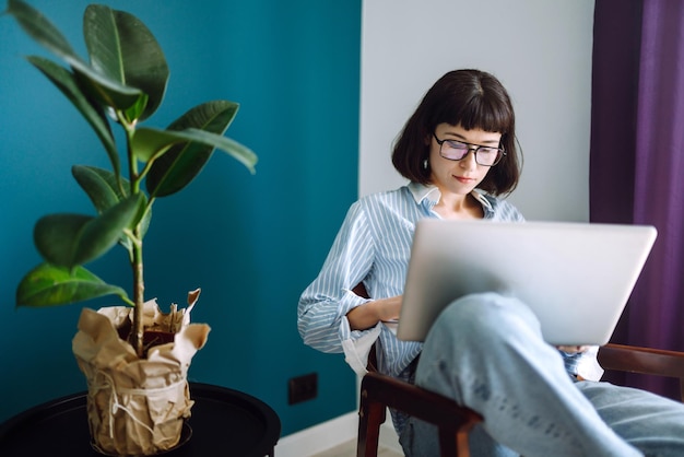 Mujer joven independiente sentada en un sofá moderno y trabajando en una computadora portátil Educación en línea Trabajar desde casa