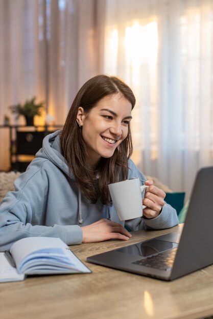 Mujer joven independiente tomando café y usando una computadora portátil en la mesa en la sala de estar de su casa