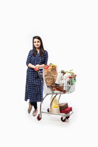 Mujer joven india con carrito de compras o carrito lleno de comestibles, verduras y frutas. Foto de cuerpo entero aislado sobre pared blanca