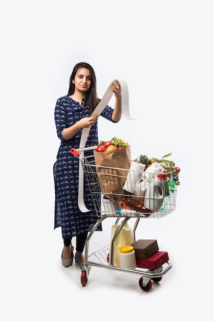 Mujer joven india con carrito de compras o carrito lleno de comestibles, verduras y frutas. Foto de cuerpo entero aislado sobre pared blanca