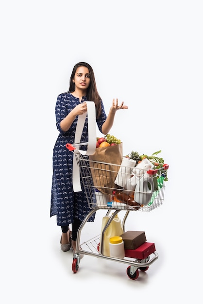 Mujer joven india con carrito de compras o carrito lleno de comestibles, verduras y frutas. Foto de cuerpo entero aislado sobre pared blanca