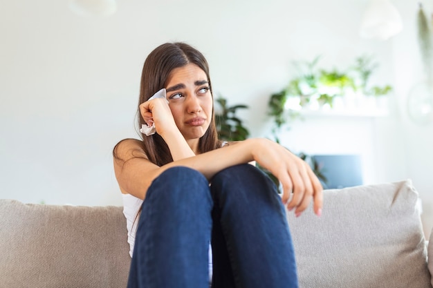 Mujer joven infeliz que cubre la cara con las manos, llorando sola de cerca, niña deprimida sentada en el sofá en casa, problema de salud o pensando en malas relaciones, romper con su novio, divorcio