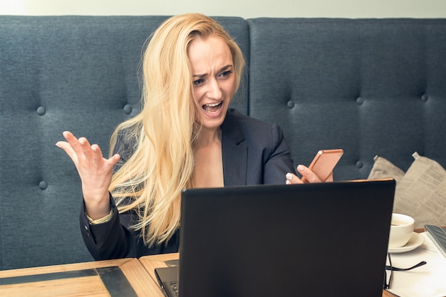 Mujer joven insatisfecha enojada que mira en el teléfono inteligente en el teléfono delante de la computadora portátil.