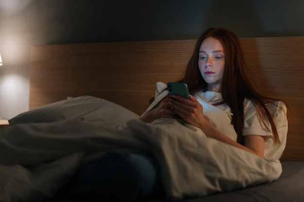 Mujer joven con insomnio grave que usa un teléfono inteligente mirando en la pantalla escribiendo un mensaje en línea en las redes sociales acostada en la cama a altas horas de la noche