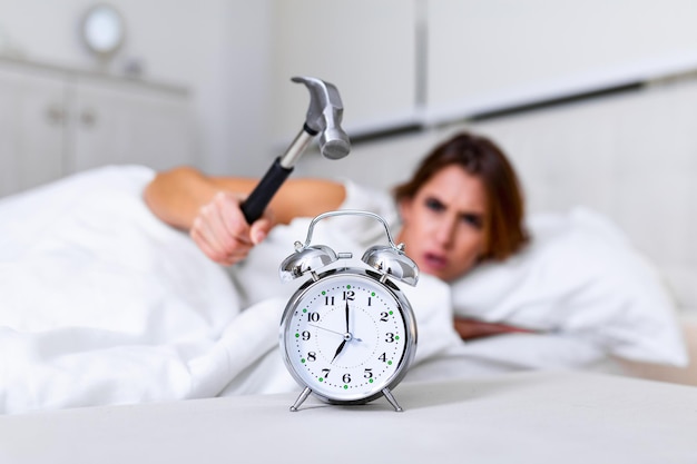 Mujer joven intenta romper el despertador con un martillo, Destroy the Clock. Chica acostada en la cama apagando un despertador con un martillo por la mañana a las 7 de la mañana.