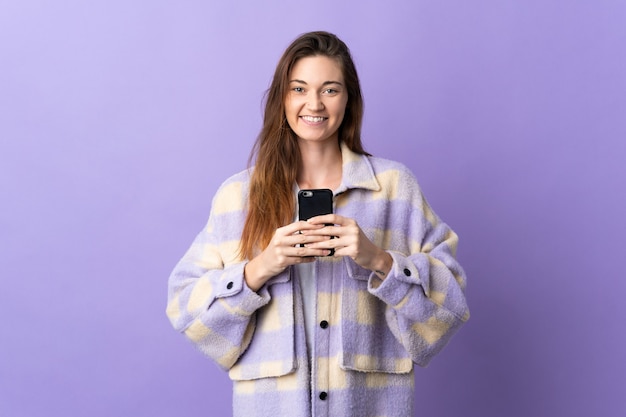 Mujer joven de Irlanda aislada sobre fondo púrpura mirando a la cámara y sonriendo mientras usa el móvil